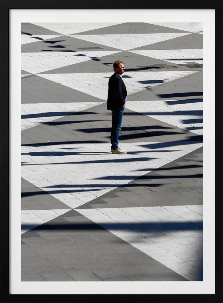 A man in a dark jacket and blue jeans stands alone in profile on a geometrically patterned plaza. The ground is a striking pattern of grey and white triangles, crossed by long, dark shadows cast by the bright sun, creating a high-contrast, graphic composition. Wall Art