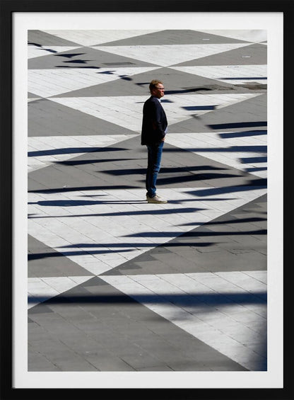 A man in a dark jacket and blue jeans stands alone in profile on a geometrically patterned plaza. The ground is a striking pattern of grey and white triangles, crossed by long, dark shadows cast by the bright sun, creating a high-contrast, graphic composition. Wall Art