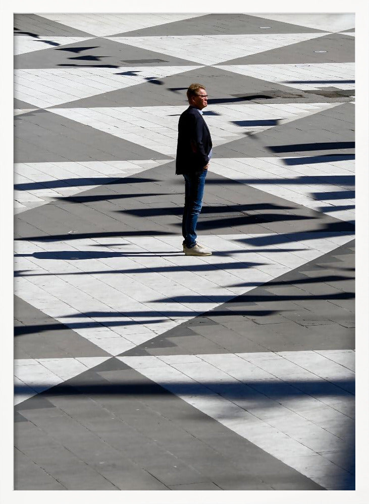 A man in a dark jacket and blue jeans stands alone in profile on a geometrically patterned plaza. The ground is a striking pattern of grey and white triangles, crossed by long, dark shadows cast by the bright sun, creating a high-contrast, graphic composition. Wall Art