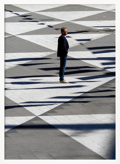 A man in a dark jacket and blue jeans stands alone in profile on a geometrically patterned plaza. The ground is a striking pattern of grey and white triangles, crossed by long, dark shadows cast by the bright sun, creating a high-contrast, graphic composition. Wall Art