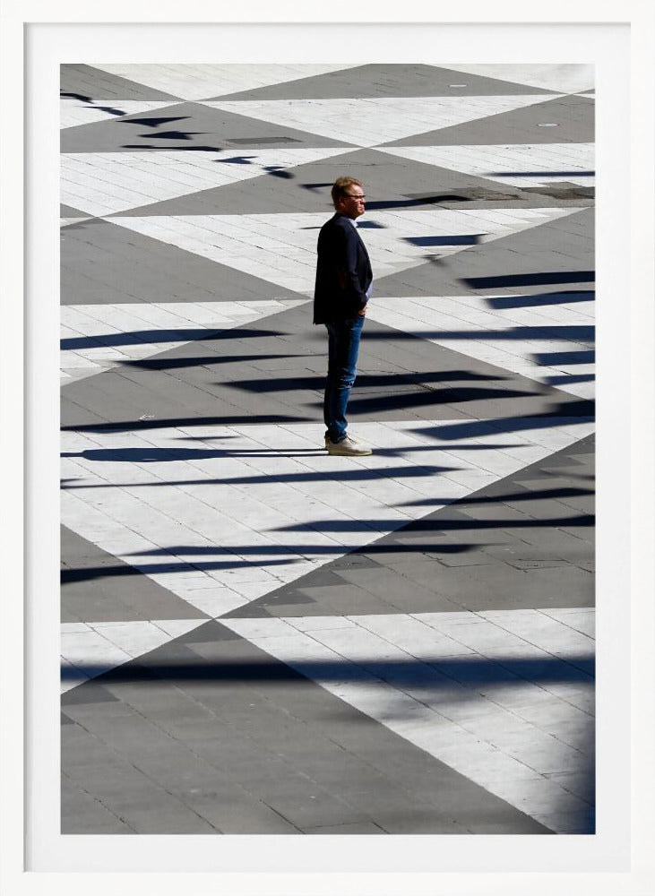 A man in a dark jacket and blue jeans stands alone in profile on a geometrically patterned plaza. The ground is a striking pattern of grey and white triangles, crossed by long, dark shadows cast by the bright sun, creating a high-contrast, graphic composition. Wall Art