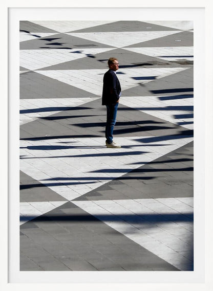 A man in a dark jacket and blue jeans stands alone in profile on a geometrically patterned plaza. The ground is a striking pattern of grey and white triangles, crossed by long, dark shadows cast by the bright sun, creating a high-contrast, graphic composition. Wall Art