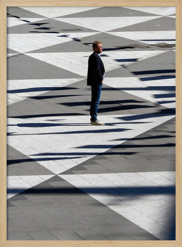 A man in a dark jacket and blue jeans stands alone in profile on a geometrically patterned plaza. The ground is a striking pattern of grey and white triangles, crossed by long, dark shadows cast by the bright sun, creating a high-contrast, graphic composition. Wall Art