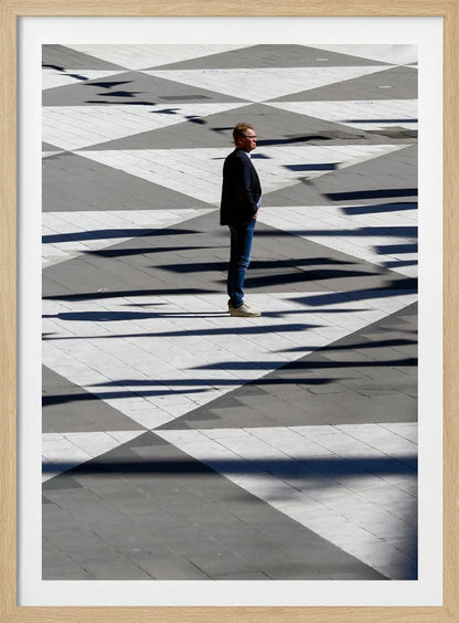 A man in a dark jacket and blue jeans stands alone in profile on a geometrically patterned plaza. The ground is a striking pattern of grey and white triangles, crossed by long, dark shadows cast by the bright sun, creating a high-contrast, graphic composition. Wall Art