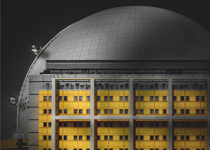 A framed architectural photograph with selective color, featuring a bright yellow building in the foreground against a monochrome background of a large grey dome structure under a black sky. Wall Art