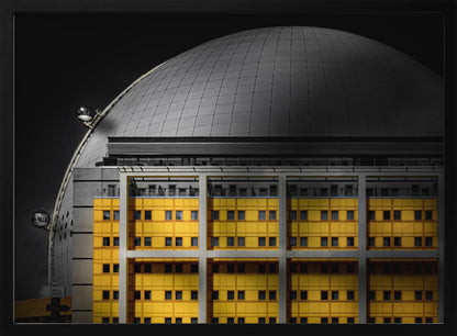 A framed architectural photograph with selective color, featuring a bright yellow building in the foreground against a monochrome background of a large grey dome structure under a black sky. Wall Art