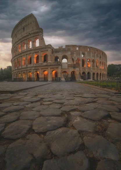 A low-angle view of the Roman Colosseum at dusk, with its arched windows glowing with warm orange light under a dramatic, stormy sky. An ancient cobblestone road leads towards the iconic amphitheater. Print