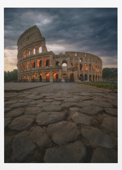 A low-angle view of the Roman Colosseum at dusk, with its arched windows glowing with warm orange light under a dramatic, stormy sky. An ancient cobblestone road leads towards the iconic amphitheater. Print