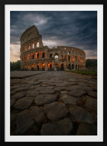 A low-angle view of the Roman Colosseum at dusk, with its arched windows glowing with warm orange light under a dramatic, stormy sky. An ancient cobblestone road leads towards the iconic amphitheater. Print