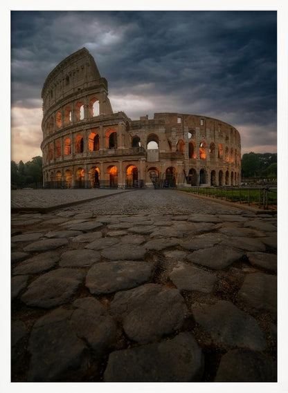 A low-angle view of the Roman Colosseum at dusk, with its arched windows glowing with warm orange light under a dramatic, stormy sky. An ancient cobblestone road leads towards the iconic amphitheater. Print