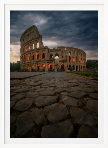 A low-angle view of the Roman Colosseum at dusk, with its arched windows glowing with warm orange light under a dramatic, stormy sky. An ancient cobblestone road leads towards the iconic amphitheater. Print