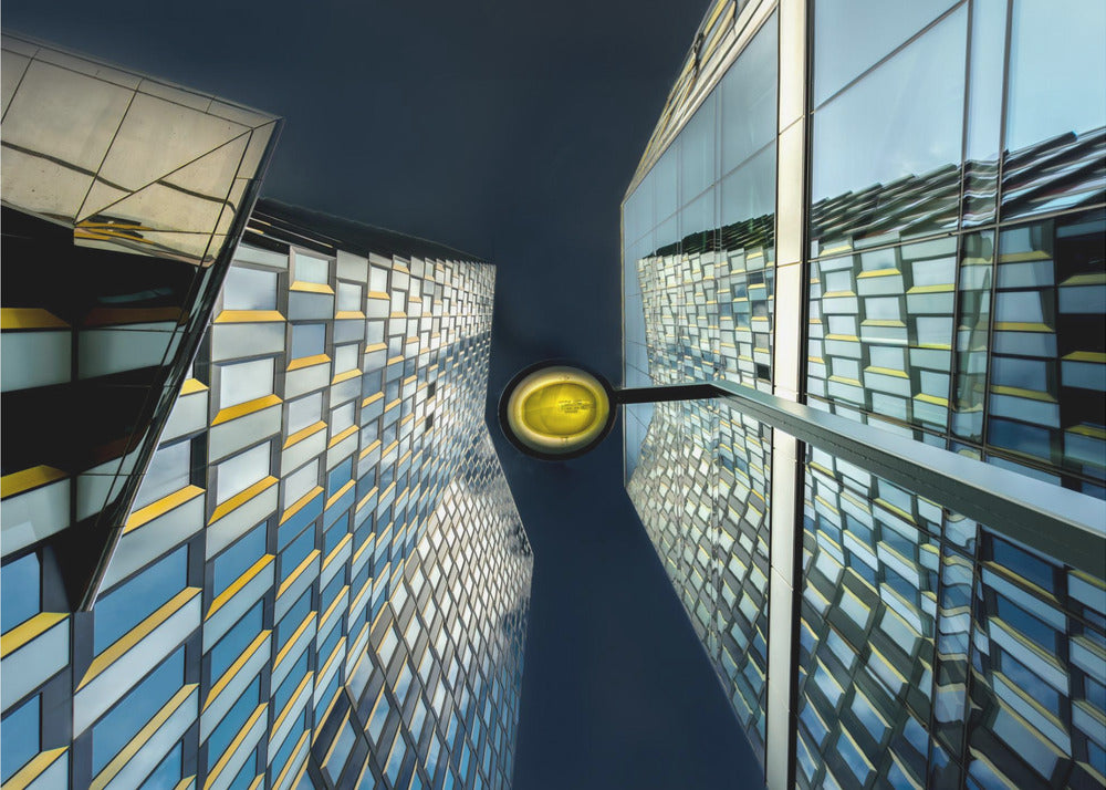 A worm's-eye view looking up between two modern glass skyscrapers with geometric blue, yellow, and silver panels, creating a symmetrical reflection against a dark blue sky, with a bright yellow streetlamp centered in the gap. Decor