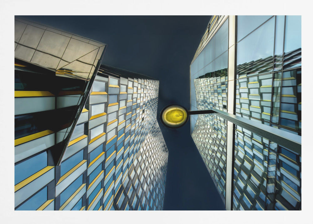 A worm's-eye view looking up between two modern glass skyscrapers with geometric blue, yellow, and silver panels, creating a symmetrical reflection against a dark blue sky, with a bright yellow streetlamp centered in the gap. Decor