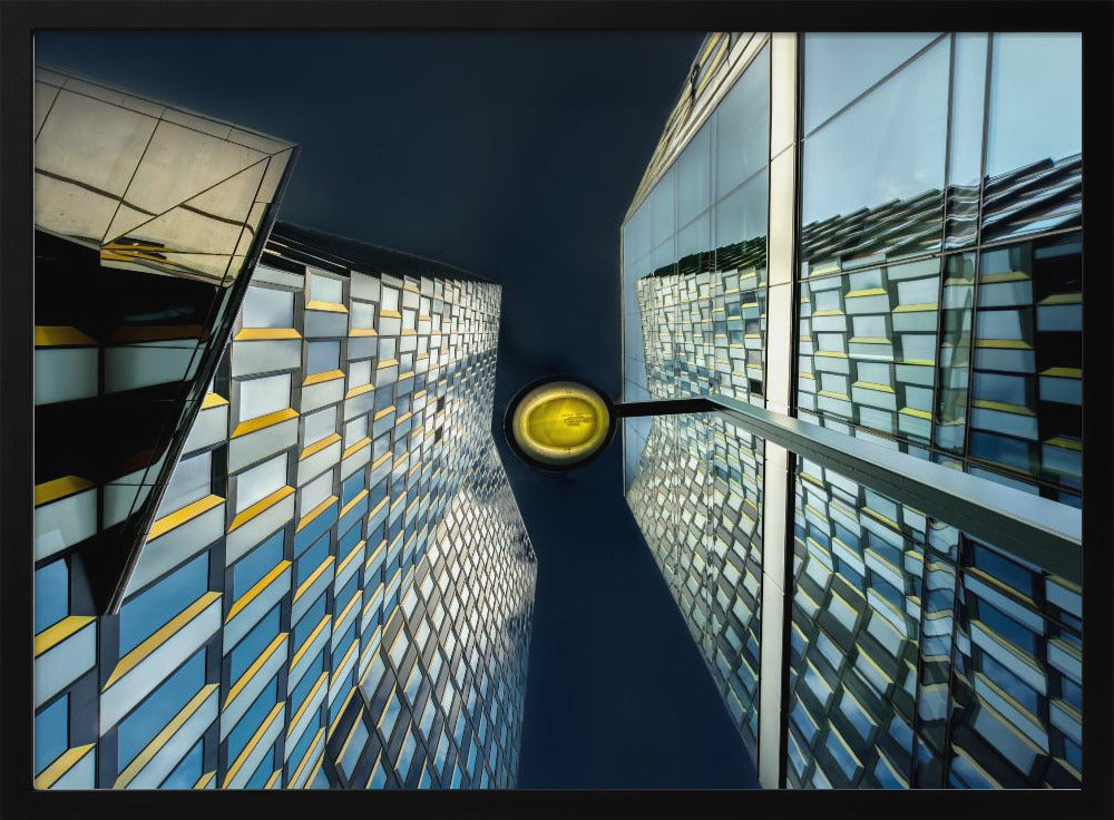 A worm's-eye view looking up between two modern glass skyscrapers with geometric blue, yellow, and silver panels, creating a symmetrical reflection against a dark blue sky, with a bright yellow streetlamp centered in the gap. Decor