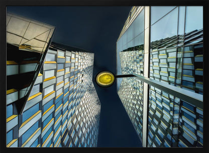 A worm's-eye view looking up between two modern glass skyscrapers with geometric blue, yellow, and silver panels, creating a symmetrical reflection against a dark blue sky, with a bright yellow streetlamp centered in the gap. Decor
