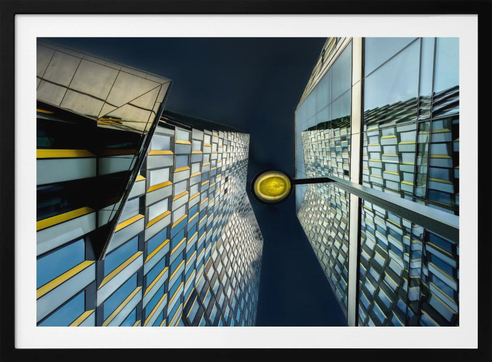 A worm's-eye view looking up between two modern glass skyscrapers with geometric blue, yellow, and silver panels, creating a symmetrical reflection against a dark blue sky, with a bright yellow streetlamp centered in the gap. Decor