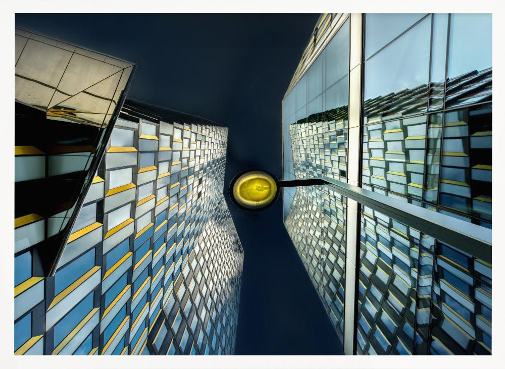 A worm's-eye view looking up between two modern glass skyscrapers with geometric blue, yellow, and silver panels, creating a symmetrical reflection against a dark blue sky, with a bright yellow streetlamp centered in the gap. Decor