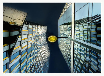 A worm's-eye view looking up between two modern glass skyscrapers with geometric blue, yellow, and silver panels, creating a symmetrical reflection against a dark blue sky, with a bright yellow streetlamp centered in the gap. Decor