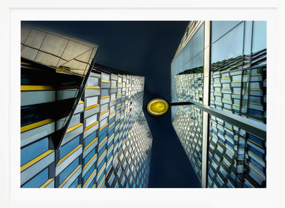 A worm's-eye view looking up between two modern glass skyscrapers with geometric blue, yellow, and silver panels, creating a symmetrical reflection against a dark blue sky, with a bright yellow streetlamp centered in the gap. Decor