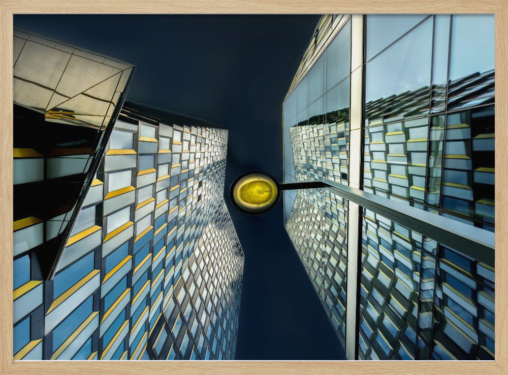 A worm's-eye view looking up between two modern glass skyscrapers with geometric blue, yellow, and silver panels, creating a symmetrical reflection against a dark blue sky, with a bright yellow streetlamp centered in the gap. Decor