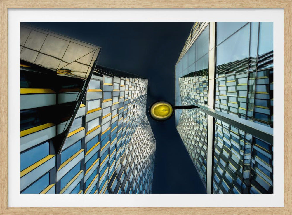 A worm's-eye view looking up between two modern glass skyscrapers with geometric blue, yellow, and silver panels, creating a symmetrical reflection against a dark blue sky, with a bright yellow streetlamp centered in the gap. Decor