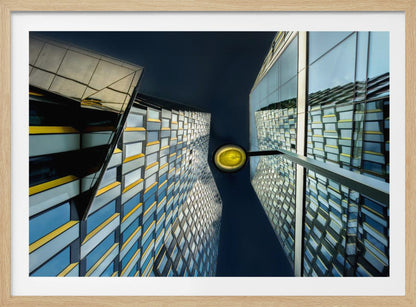 A worm's-eye view looking up between two modern glass skyscrapers with geometric blue, yellow, and silver panels, creating a symmetrical reflection against a dark blue sky, with a bright yellow streetlamp centered in the gap. Decor