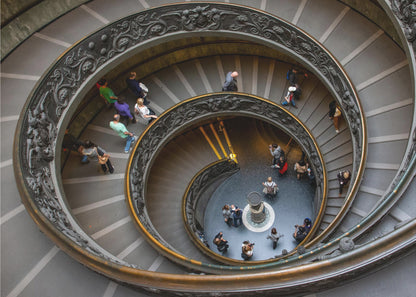 A framed, high-angle photograph looking down into the center of a grand spiral staircase. The gray stone steps spiral downwards, flanked by ornate, dark bronze railings. Numerous people are scattered along the stairs, appearing small from the overhead perspective, giving a sense of the structure's massive scale. Artwork