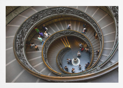 A framed, high-angle photograph looking down into the center of a grand spiral staircase. The gray stone steps spiral downwards, flanked by ornate, dark bronze railings. Numerous people are scattered along the stairs, appearing small from the overhead perspective, giving a sense of the structure's massive scale. Artwork