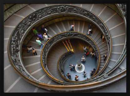 A framed, high-angle photograph looking down into the center of a grand spiral staircase. The gray stone steps spiral downwards, flanked by ornate, dark bronze railings. Numerous people are scattered along the stairs, appearing small from the overhead perspective, giving a sense of the structure's massive scale. Artwork
