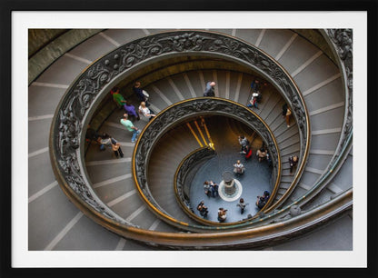 A framed, high-angle photograph looking down into the center of a grand spiral staircase. The gray stone steps spiral downwards, flanked by ornate, dark bronze railings. Numerous people are scattered along the stairs, appearing small from the overhead perspective, giving a sense of the structure's massive scale. Artwork