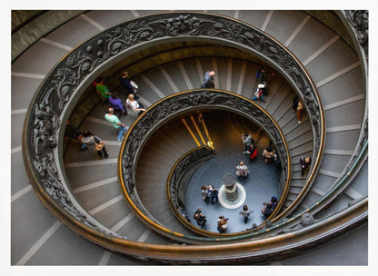 A framed, high-angle photograph looking down into the center of a grand spiral staircase. The gray stone steps spiral downwards, flanked by ornate, dark bronze railings. Numerous people are scattered along the stairs, appearing small from the overhead perspective, giving a sense of the structure's massive scale. Artwork