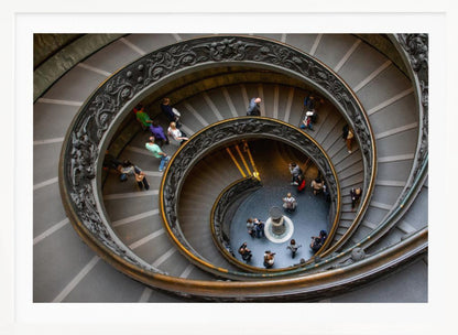 A framed, high-angle photograph looking down into the center of a grand spiral staircase. The gray stone steps spiral downwards, flanked by ornate, dark bronze railings. Numerous people are scattered along the stairs, appearing small from the overhead perspective, giving a sense of the structure's massive scale. Artwork