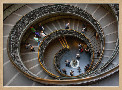 A framed, high-angle photograph looking down into the center of a grand spiral staircase. The gray stone steps spiral downwards, flanked by ornate, dark bronze railings. Numerous people are scattered along the stairs, appearing small from the overhead perspective, giving a sense of the structure's massive scale. Artwork