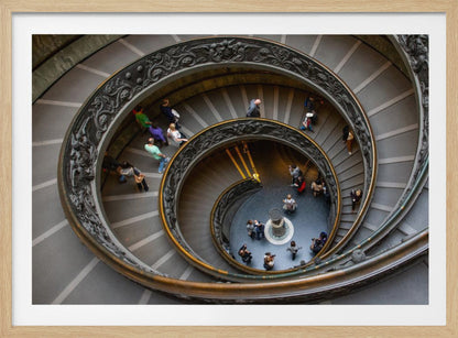 A framed, high-angle photograph looking down into the center of a grand spiral staircase. The gray stone steps spiral downwards, flanked by ornate, dark bronze railings. Numerous people are scattered along the stairs, appearing small from the overhead perspective, giving a sense of the structure's massive scale. Artwork