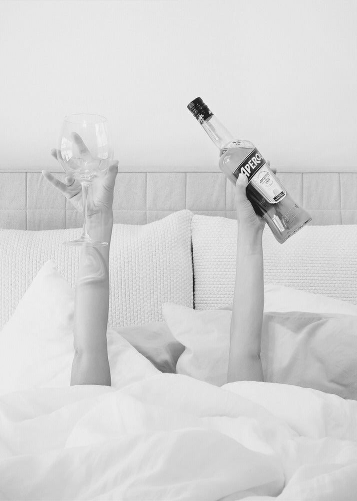 A black and white photograph of a person under white covers in bed, with only their arms visible. One hand holds up a bottle of Aperol, and the other holds up an empty wine glass. Decor