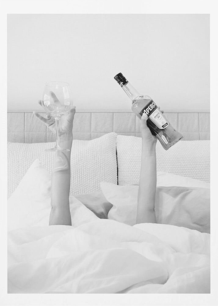 A black and white photograph of a person under white covers in bed, with only their arms visible. One hand holds up a bottle of Aperol, and the other holds up an empty wine glass. Decor