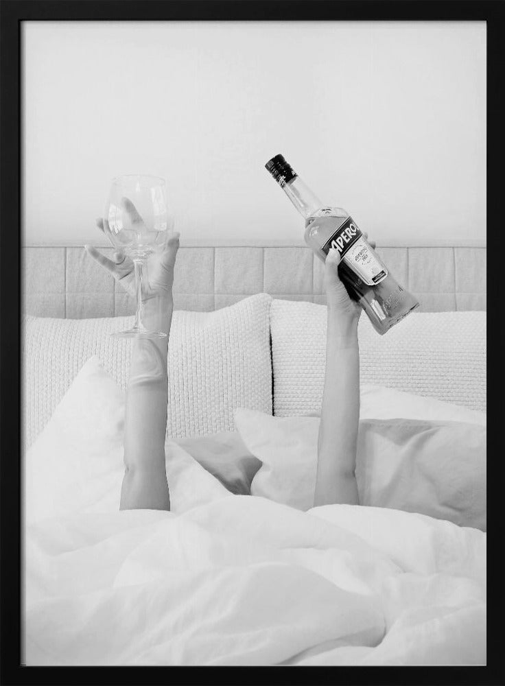 A black and white photograph of a person under white covers in bed, with only their arms visible. One hand holds up a bottle of Aperol, and the other holds up an empty wine glass. Decor