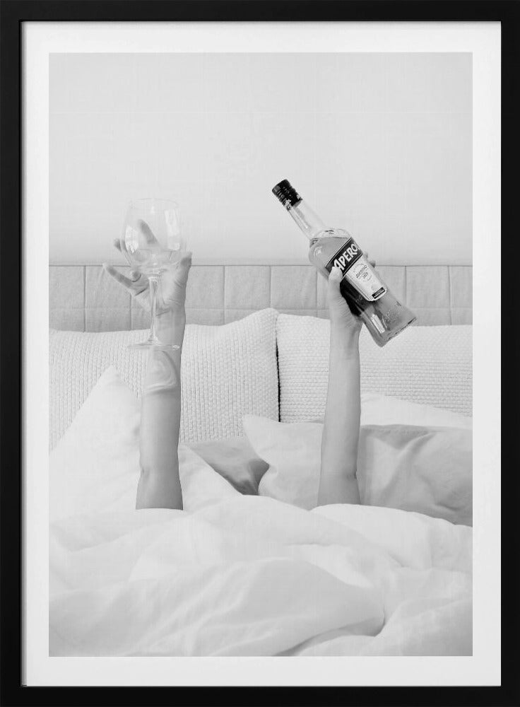 A black and white photograph of a person under white covers in bed, with only their arms visible. One hand holds up a bottle of Aperol, and the other holds up an empty wine glass. Decor