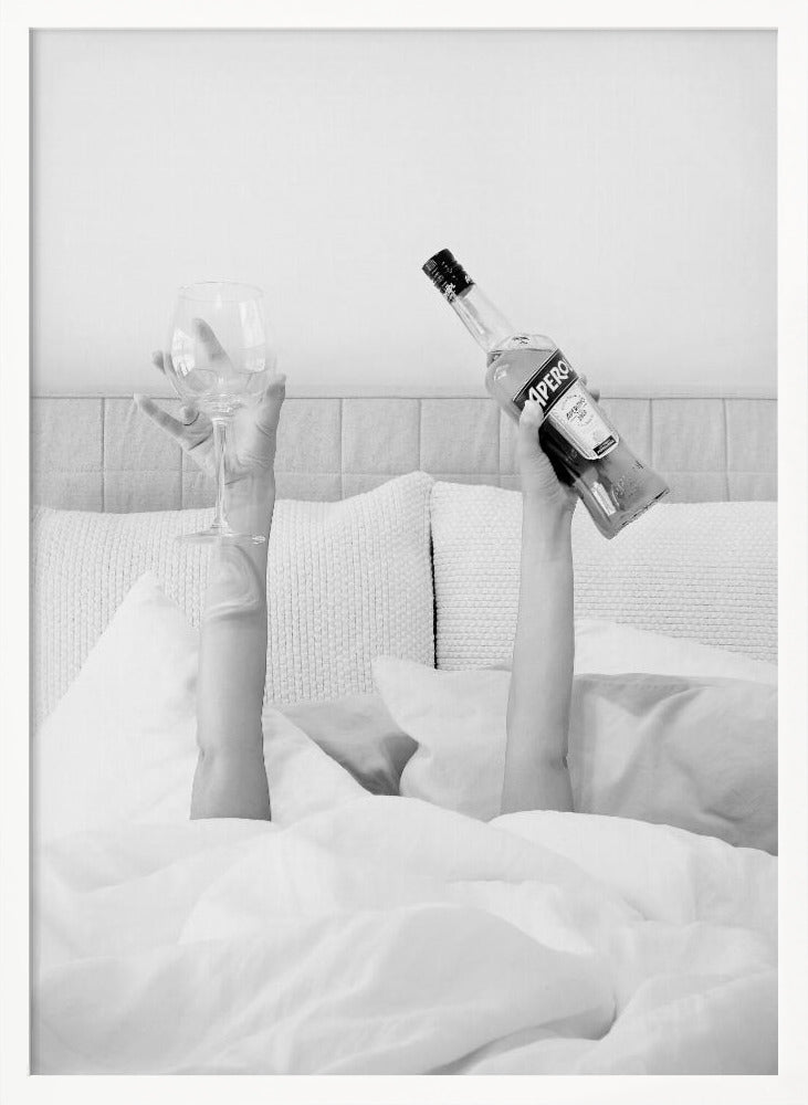 A black and white photograph of a person under white covers in bed, with only their arms visible. One hand holds up a bottle of Aperol, and the other holds up an empty wine glass. Decor
