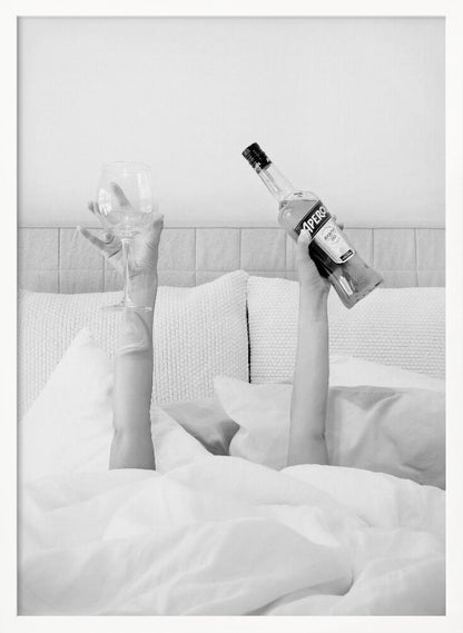 A black and white photograph of a person under white covers in bed, with only their arms visible. One hand holds up a bottle of Aperol, and the other holds up an empty wine glass. Decor