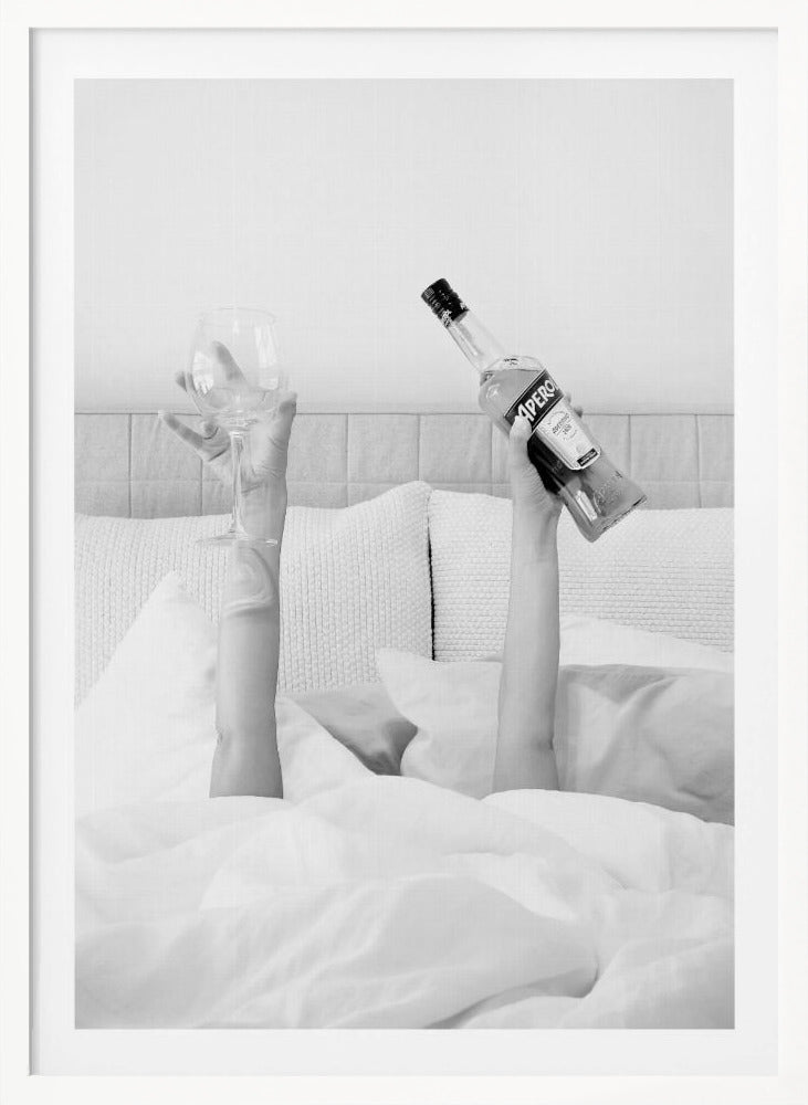 A black and white photograph of a person under white covers in bed, with only their arms visible. One hand holds up a bottle of Aperol, and the other holds up an empty wine glass. Decor