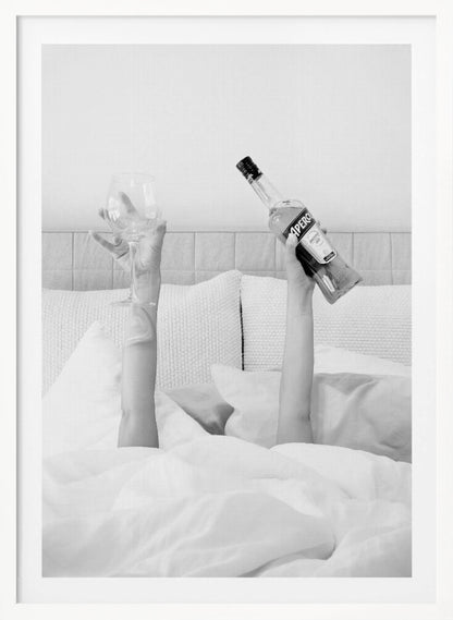 A black and white photograph of a person under white covers in bed, with only their arms visible. One hand holds up a bottle of Aperol, and the other holds up an empty wine glass. Decor