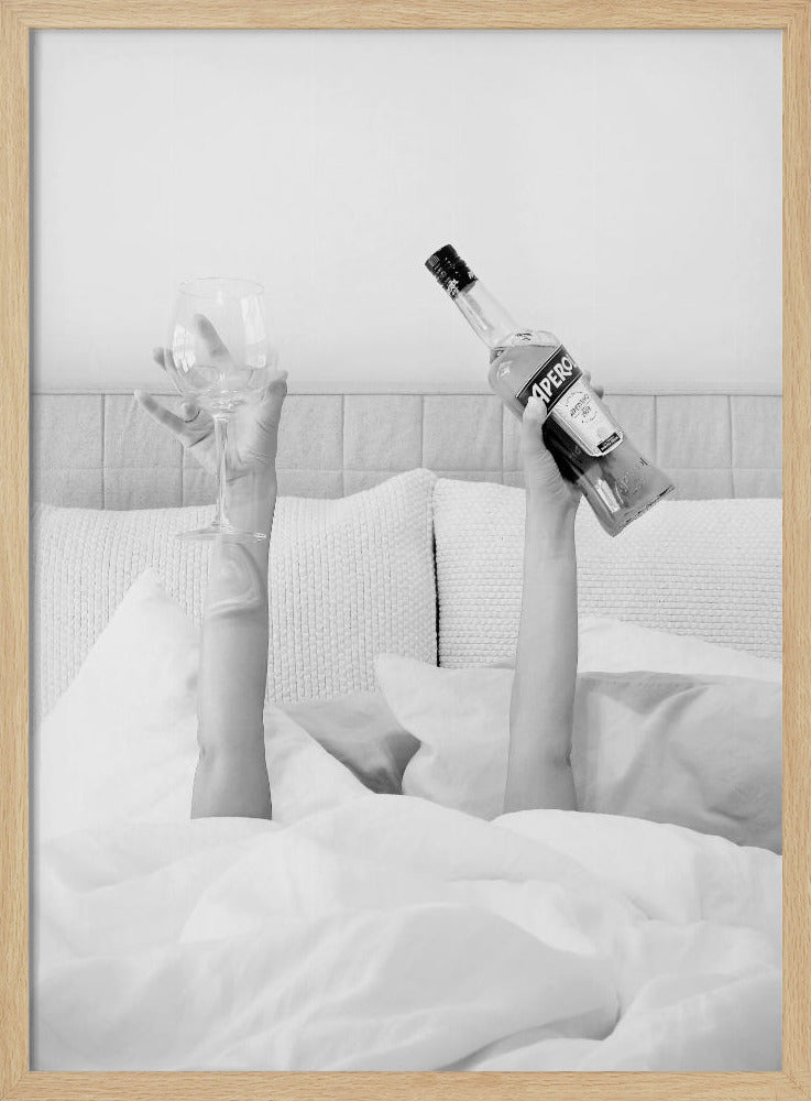 A black and white photograph of a person under white covers in bed, with only their arms visible. One hand holds up a bottle of Aperol, and the other holds up an empty wine glass. Decor
