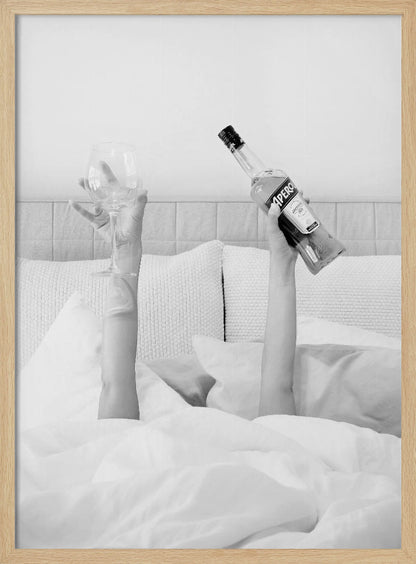 A black and white photograph of a person under white covers in bed, with only their arms visible. One hand holds up a bottle of Aperol, and the other holds up an empty wine glass. Decor