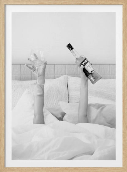 A black and white photograph of a person under white covers in bed, with only their arms visible. One hand holds up a bottle of Aperol, and the other holds up an empty wine glass. Decor