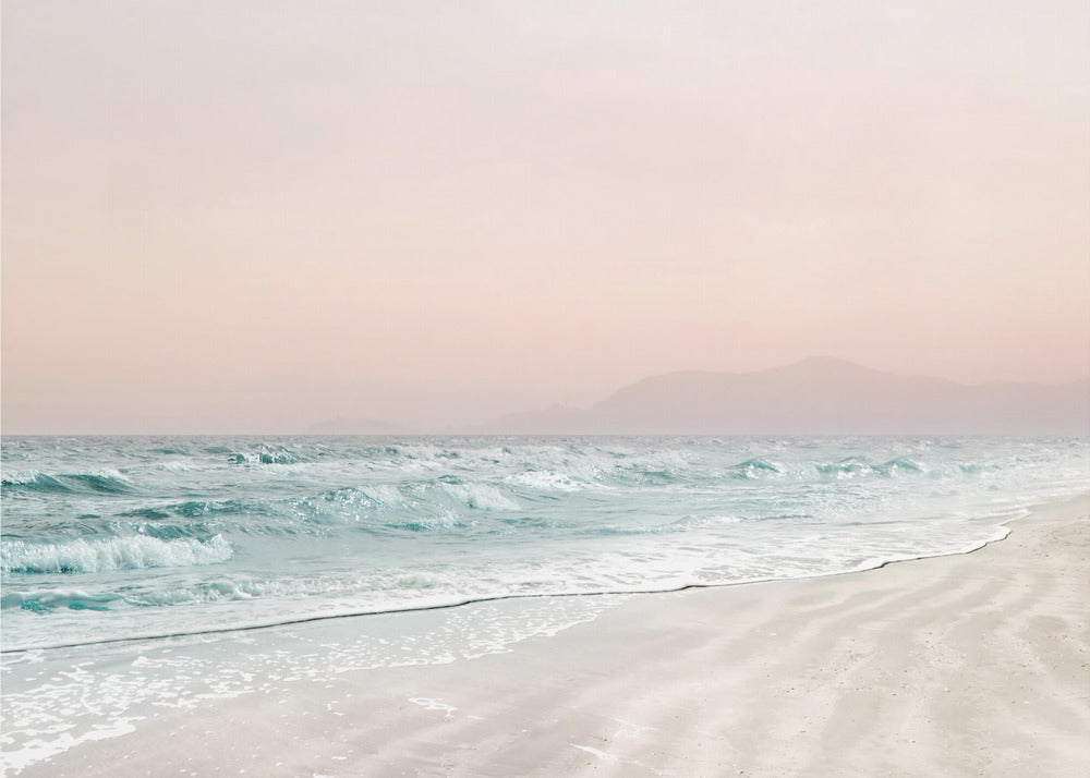 A serene coastal scene with gentle turquoise waves washing onto a sandy beach. The sky is a soft, hazy pink, and faint outlines of mountains are visible in the distant background. The artwork is presented in a silver frame. Print