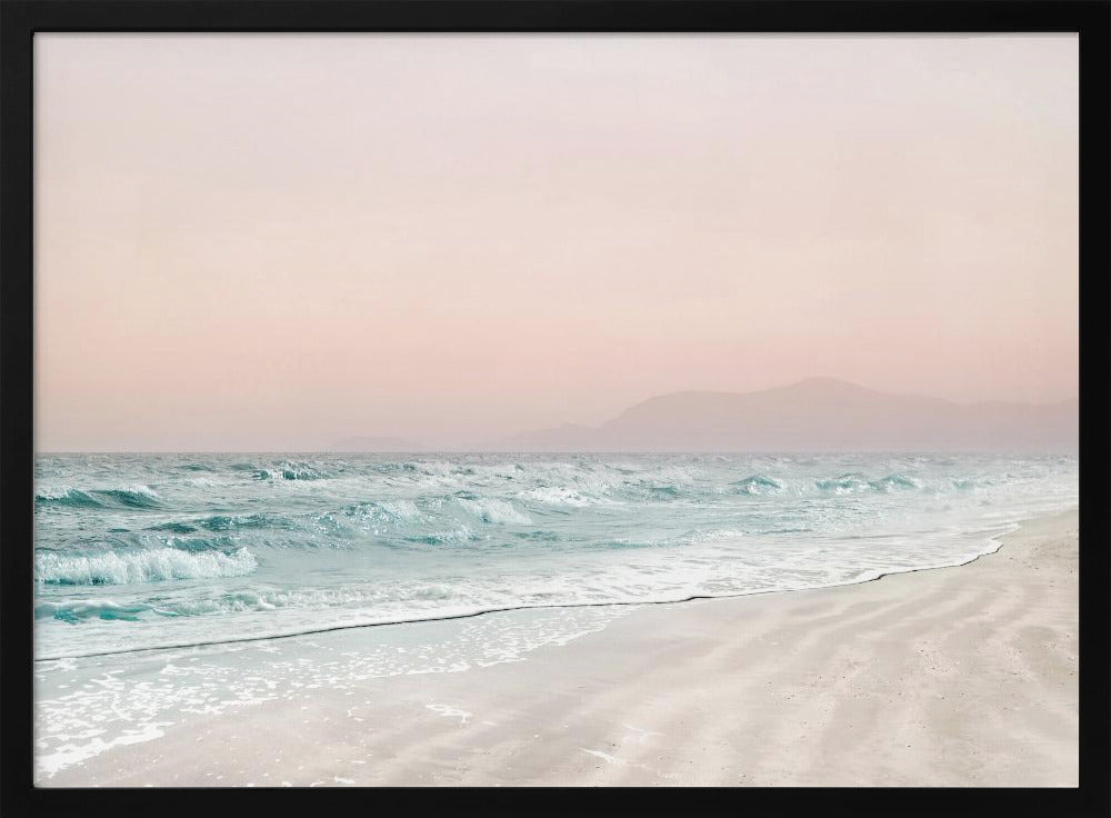 A serene coastal scene with gentle turquoise waves washing onto a sandy beach. The sky is a soft, hazy pink, and faint outlines of mountains are visible in the distant background. The artwork is presented in a silver frame. Print