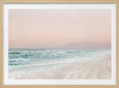 A serene coastal scene with gentle turquoise waves washing onto a sandy beach. The sky is a soft, hazy pink, and faint outlines of mountains are visible in the distant background. The artwork is presented in a silver frame. Print