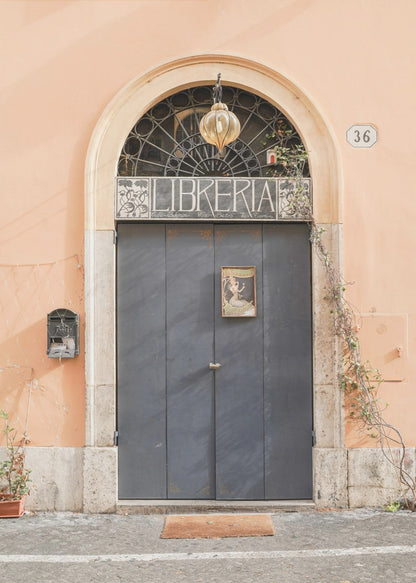 A charming European bookstore entrance featuring a large, dark grey arched door set against a peach-colored wall. Above the door, a sign reads 'LIBRERIA' below a decorative fanlight window and a hanging lantern. Decor