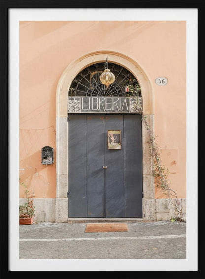 A charming European bookstore entrance featuring a large, dark grey arched door set against a peach-colored wall. Above the door, a sign reads 'LIBRERIA' below a decorative fanlight window and a hanging lantern. Decor